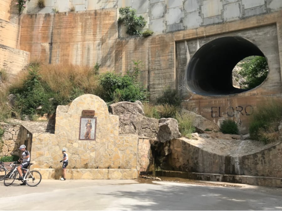 A large hole for water drainage, towers over cyclists at the water fountain below.